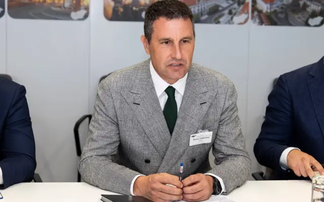 Barna Tanczos, fostul ministru, este asezat la o masa intr-un cadru oficial, purtand un sacou in carouri si o cravata verde