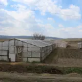 Two greenhouses on a small farm with a cultivated field and people walking in the distance