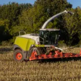 O combină agricolă modernă, de culoare verde deschis și alb, cu un cap de recoltare roșu, lucrează pe un câmp de porumb deja recoltat, lăsând în urmă miriște
