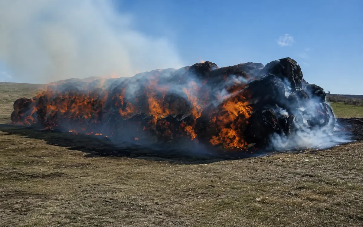 Large stack of hay bales engulfed in intense orange flames and thick smoke in an open field