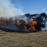Large stack of hay bales engulfed in intense orange flames and thick smoke in an open field