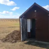 A small wooden shed housing equipment for water drilling on a farm field under a blue sky