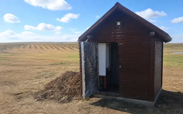 A small wooden shed housing equipment for water drilling on a farm field under a blue sky