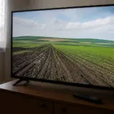 Un televizor cu ecran mare așezat pe o comodă de lemn, difuzând o imagine panoramică a unui câmp agricol cu rânduri cultivate, sub un cer cu nori